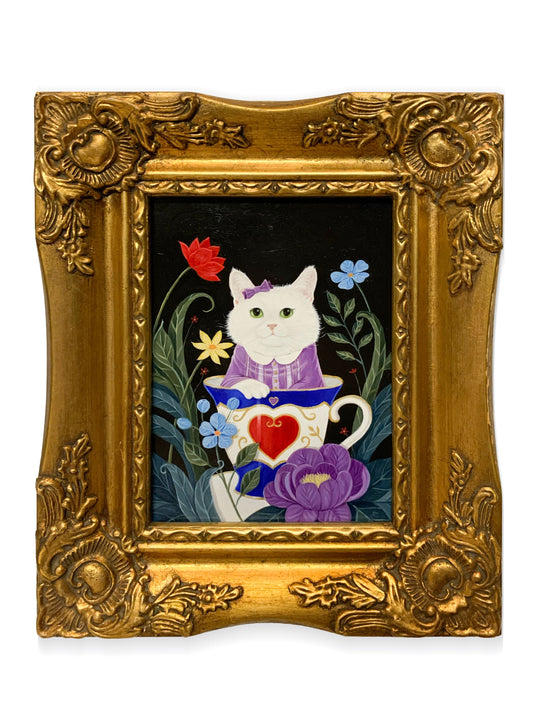 A framed art work featuring a white cat in a purple dress sitting in a teacup with flowers and green foliage on a black background, enclosed in an ornate gold frame.