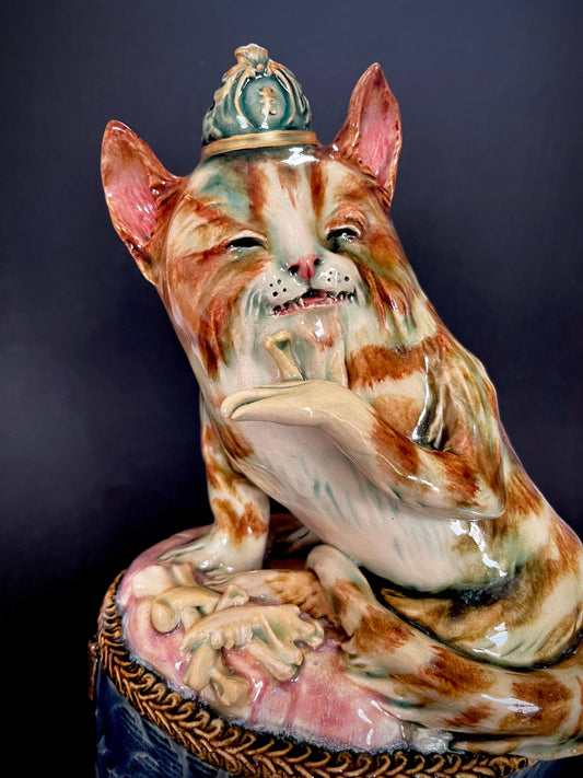 A glossy ceramic figurine of a cat wearing a crown, sitting on a decorative pedestal.