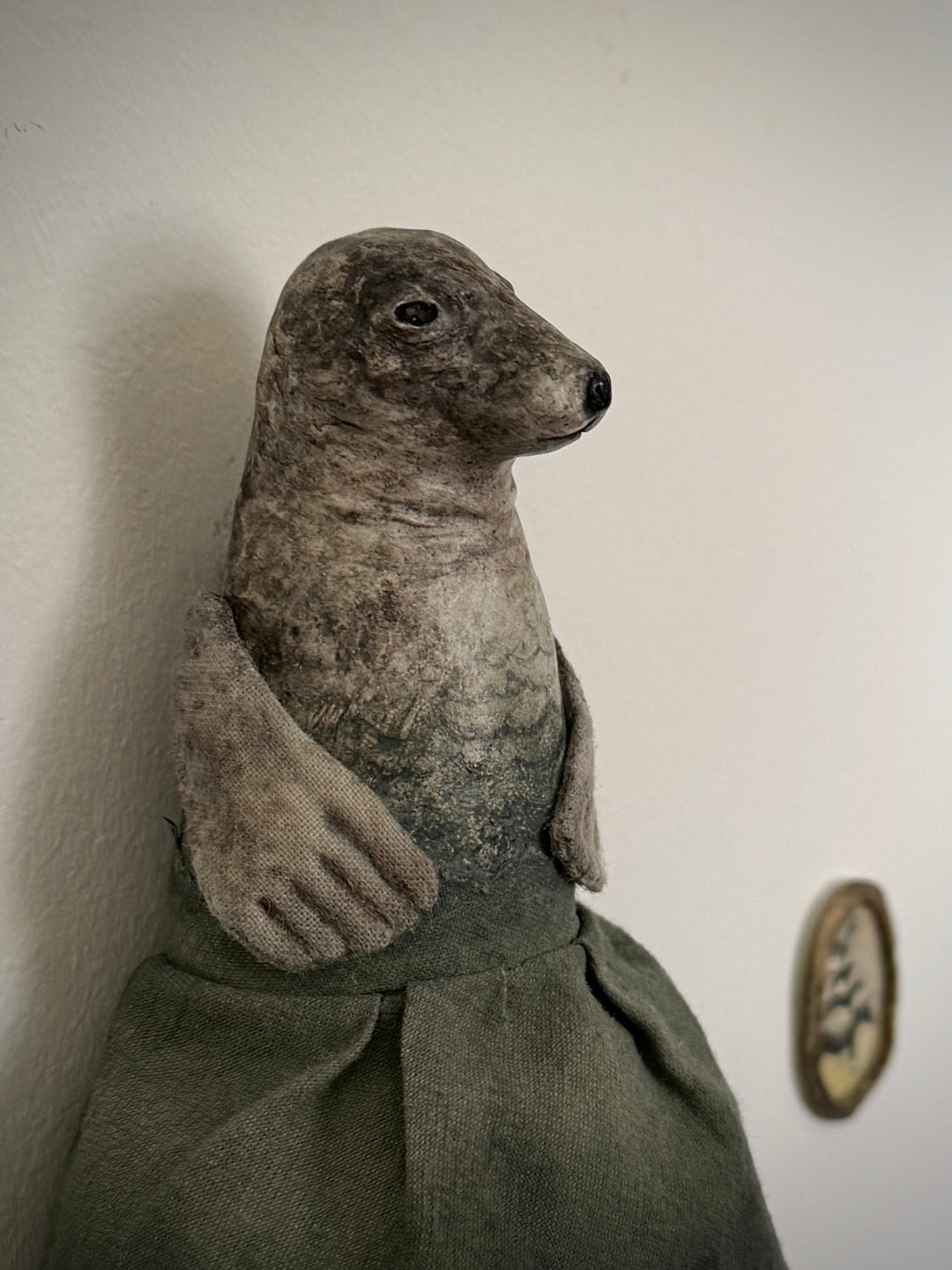 A handmade seal sculpture wearing a dark green dress, posed against a plain off-white wall.