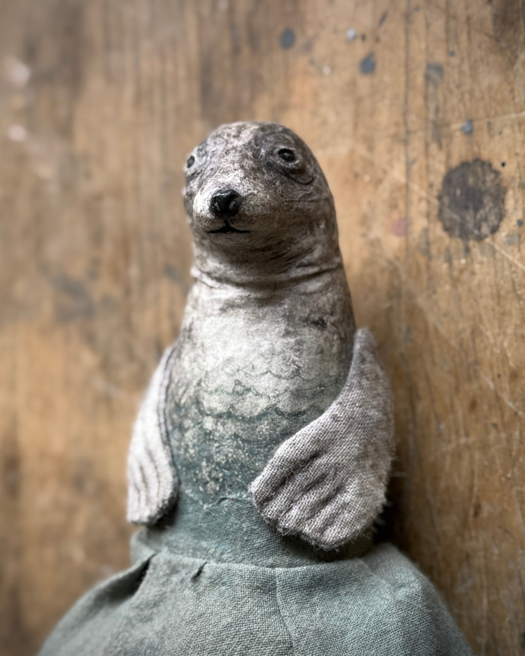 A handmade fabric seal doll with textured gray and white fur-like detailing, posed against a rustic wooden background.