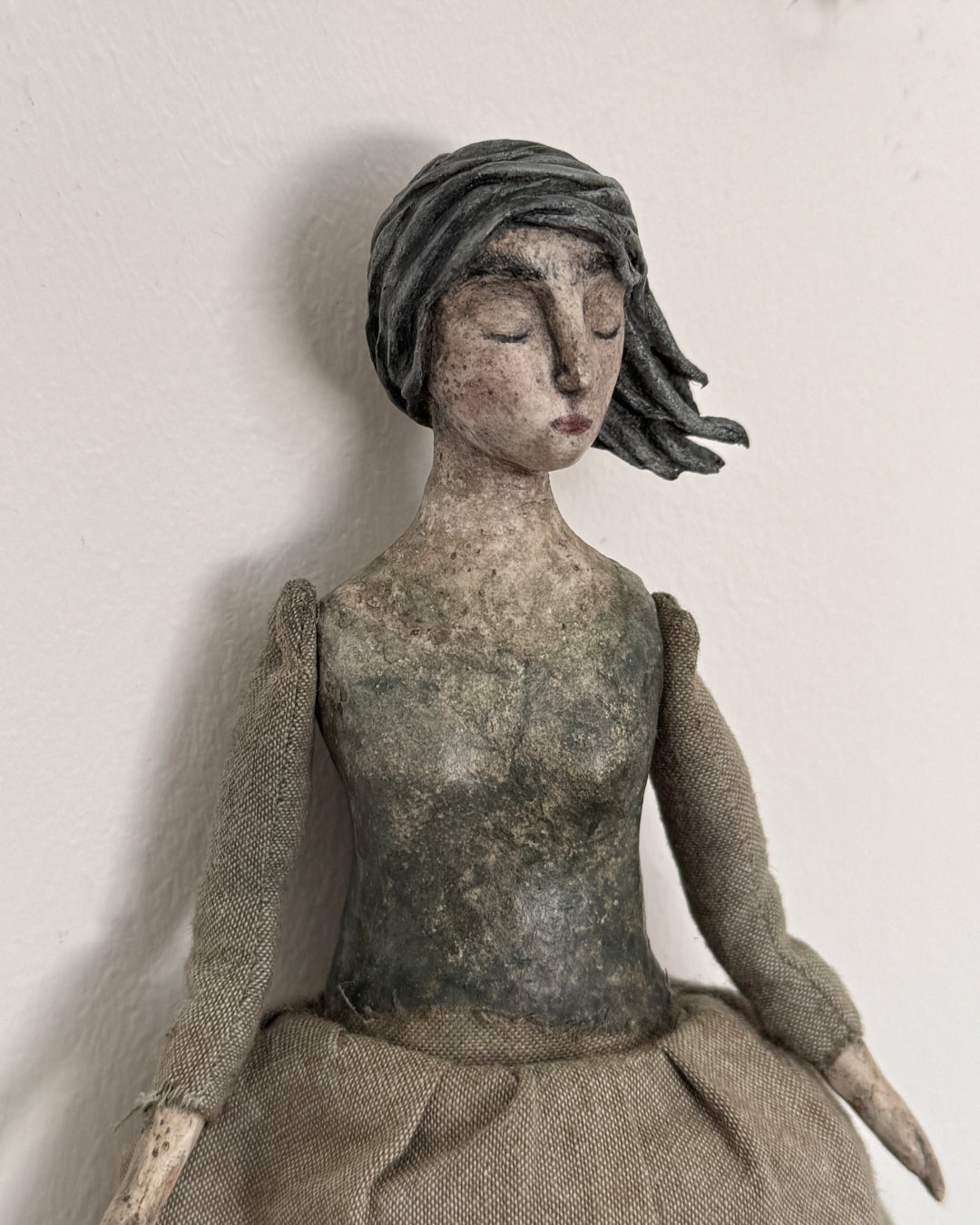 A rustic handmade doll with a weathered gray-green face and torso, wearing a textured burlap dress and dark gray hair, photographed against a plain off-white wall.