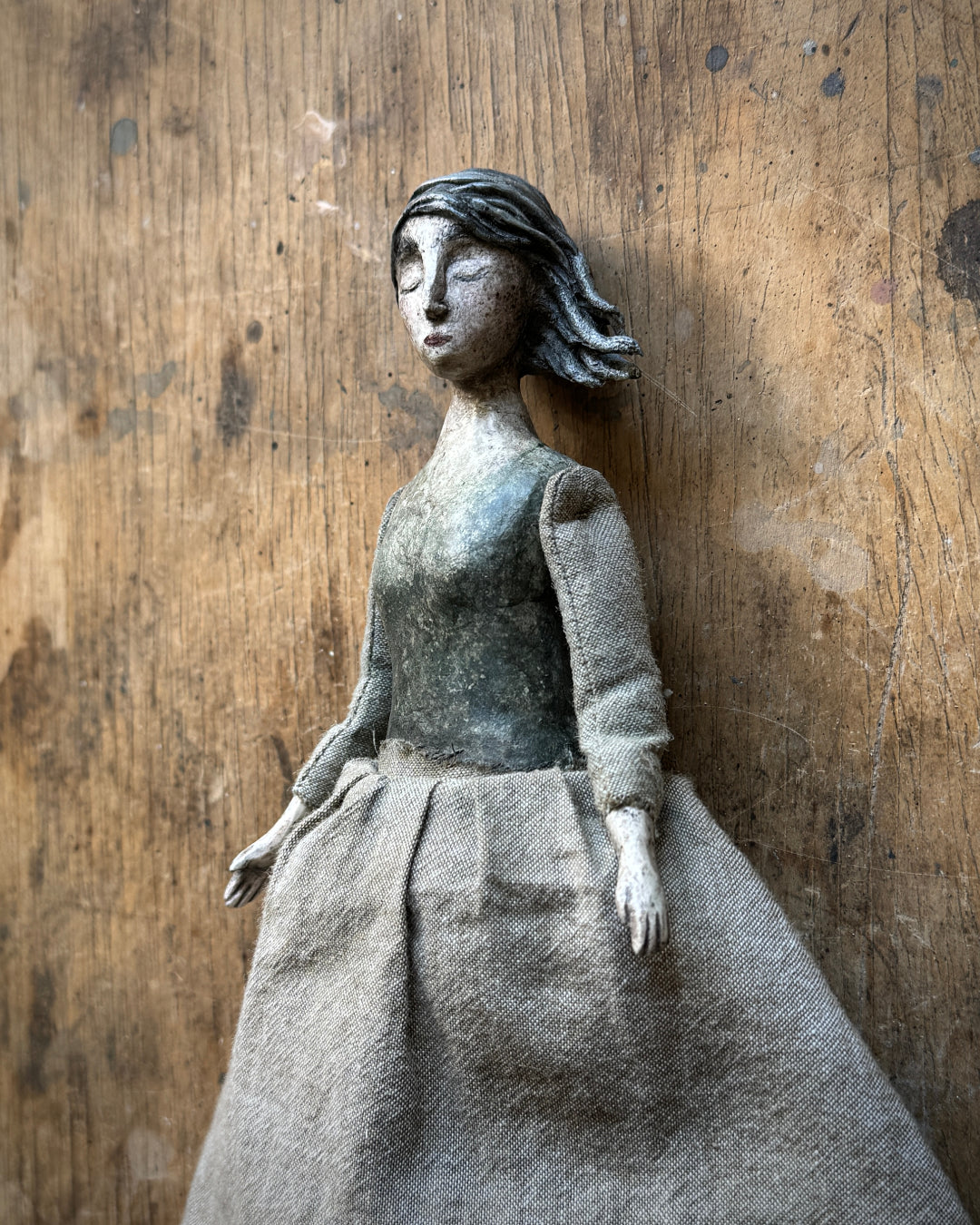 A handmade fabric doll with a weathered gray and black painted face and hair, wearing a textured gray dress, posed against a rustic wooden background.