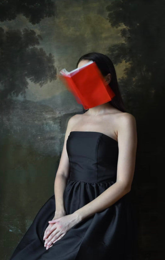 Woman in a black dress holding a red book over her face against a dark, forest-like background