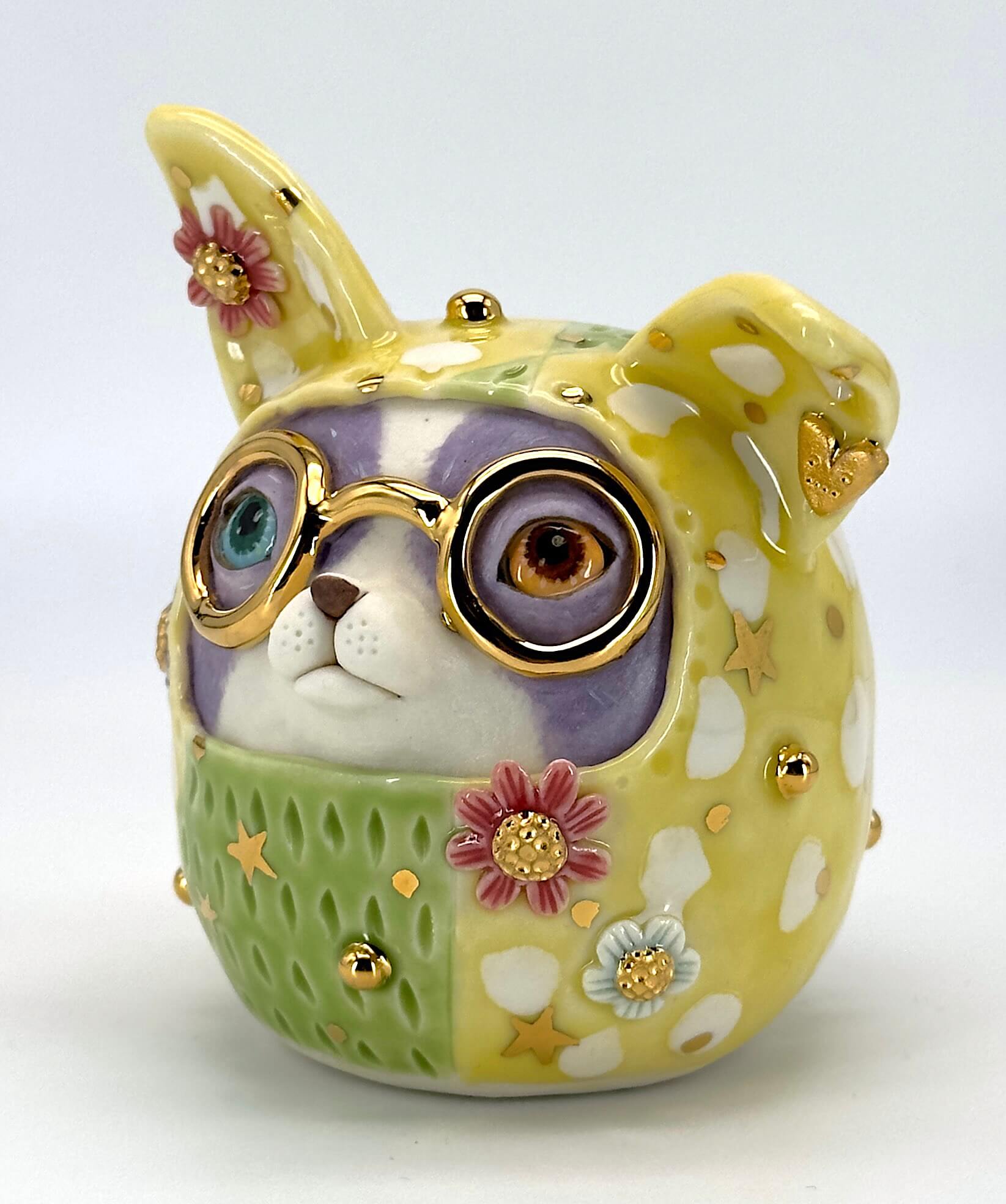 A ceramic dog-shaped sculpture featuring round gold-rimmed glasses, a lavender-purple face covering, and a green patterned lower section with yellow spots, white spots, pink flowers, and small gold stars.