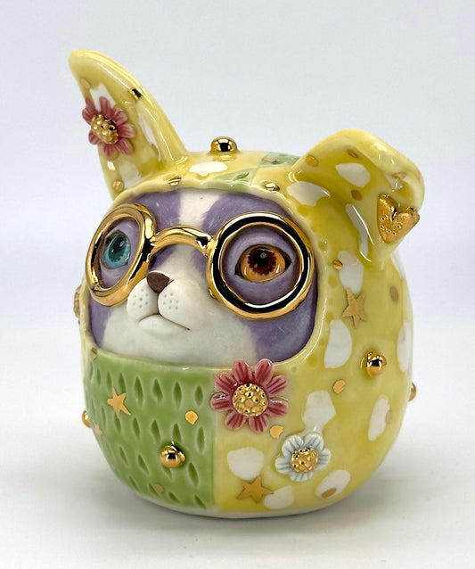 A ceramic dog-shaped sculpture featuring round gold-rimmed glasses, a lavender-purple face covering, and a green patterned lower section with yellow spots, white spots, pink flowers, and small gold stars.
