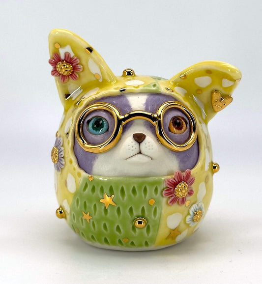 A ceramic figurine of a dog wearing round gold-rimmed glasses, a green knit sweater with gold stars, and a purple face, decorated with pink and white flowers on a light gray background.